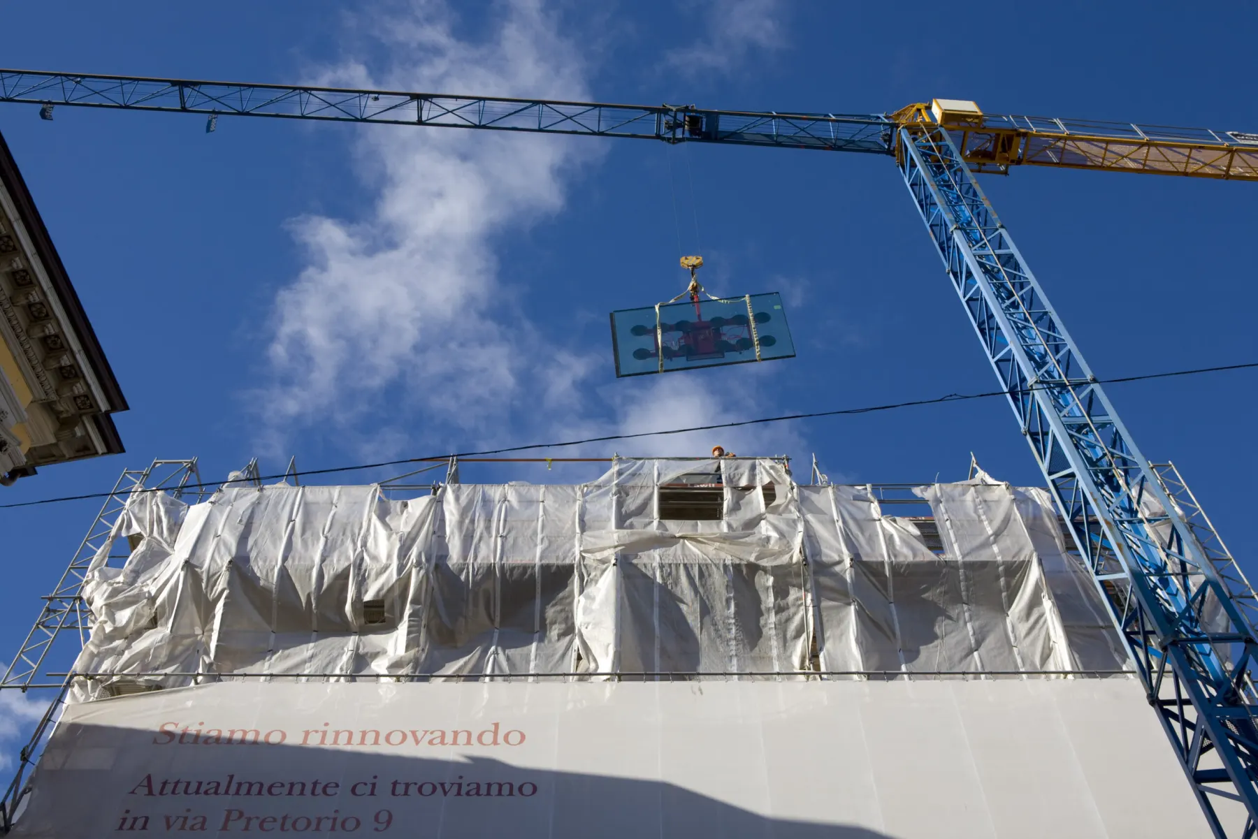 Gru al lavoro sopra uno stabile avvolto dai teli di cantiere